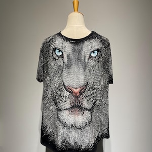 Peut inclure: Un t-shirt noir à manches courtes orné d'un grand imprimé graphique détaillé représentant un visage de lion. Le lion a des yeux bleu clair saisissants, un nez rosé et un pelage texturé gris et noir sur le devant.