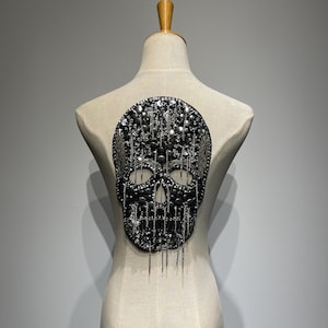 May include: A decorative skull applique, featuring a detailed design of black and silver beads and sequins. The skull is embellished with hanging chain accents, creating a gothic or punk aesthetic. The applique is displayed on a mannequin.