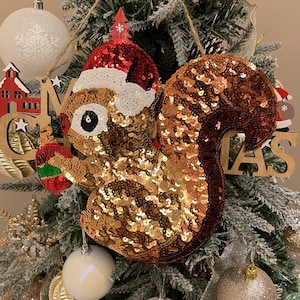 May include: A gold and brown sequined squirrel ornament with a red Santa hat. The ornament is hanging on a Christmas tree with other ornaments and a wooden sign that says "Merry Christmas".