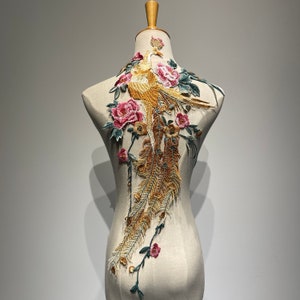 Könnte beinhalten: Eine goldene und braune Stickerei mit einem Phönix und rosa und grünen Blumen. Die Applikation befindet sich auf einem weißen Stoffgrund.