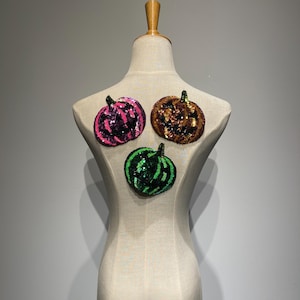 Peut inclure: Trois écussons de citrouille à paillettes roses, marron et vertes sont présentés sur un mannequin. Les citrouilles ont un aspect texturé et scintillant, chaque écusson présentant une palette de couleurs différente. Le mannequin est de couleur beige neutre.
