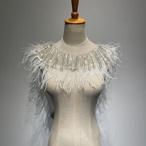 May include: A decorative white feather and sequin collar. The collar is displayed on a mannequin. The top of the collar is embellished with silver sequins, and the bottom edge is adorned with long, soft white feathers. The collar is designed to be worn around the neck.