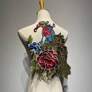 May include: Embroidered peacock and floral applique in shades of blue, red, and green. The design features a peacock with a detailed tail, a blue peony, and a red rose. The applique is displayed on a mannequin.