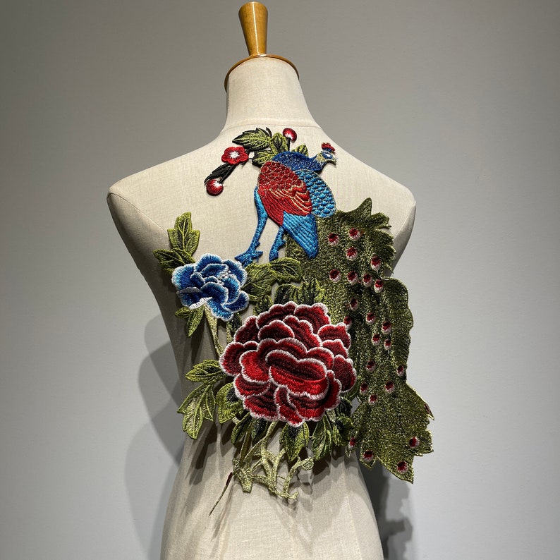 May include: Embroidered peacock and floral applique in shades of blue, red, and green. The design features a peacock with a detailed tail, a blue peony, and a red rose. The applique is displayed on a mannequin.