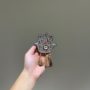 May include: A silver and black hamsa hand charm with a blue and red eye, decorated with beads and rhinestones. The charm has gold tassels hanging from the bottom.