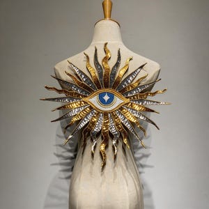 Puede incluir: Un diseño decorativo de ojo de sol con elementos metálicos dorados, plateados y bronce. El ojo presenta un iris azul con un diamante blanco. El diseño se exhibe en un maniquí.