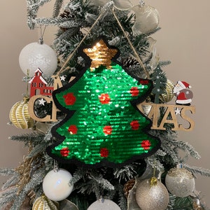 Könnte beinhalten: Ein grüner, mit Pailletten besetzter Weihnachtsbaum-Anhänger mit roten Pailletten. Der Anhänger hängt an einem Weihnachtsbaum mit anderen Anhängern und einem Holzschild mit der Aufschrift "Weihnachten".