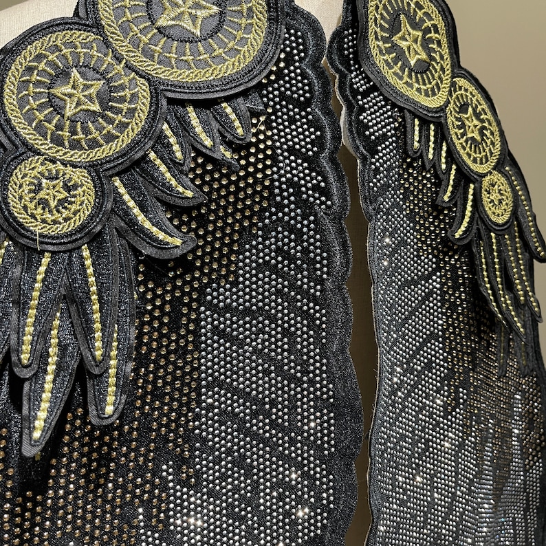 May include: Two black fabric patches with gold embroidery and rhinestones. The patches feature a wing design with a star and circle motif.