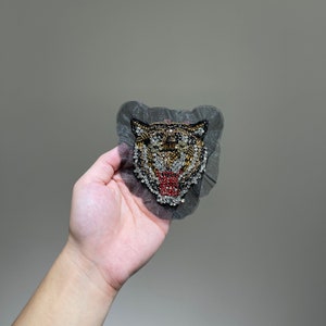 May include: A black mesh patch with a beaded tiger face design. The tiger has gold, black, and red beads for its eyes, nose, and mouth.