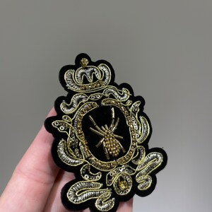 May include: A black velvet patch with gold and silver embroidery depicting a crown, a spider, and a decorative frame.