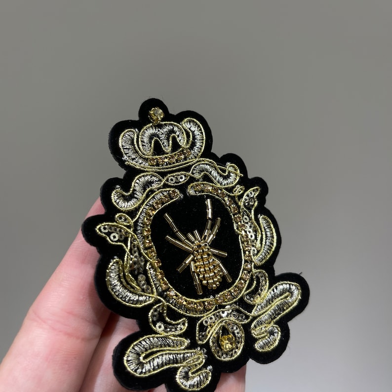 May include: A black velvet patch with gold and silver embroidery depicting a crown, a spider, and a decorative frame.