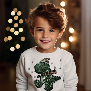 May include: A young boy wearing a white sweatshirt with a green sequined dinosaur wearing a red and white Santa hat and scarf. The dinosaur is walking and has snowflakes around it.