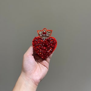 Puede incluir: Un parche bordado en forma de corazón rojo con una corona dorada y pedrería. El parche está hecho de fieltro y lentejuelas.