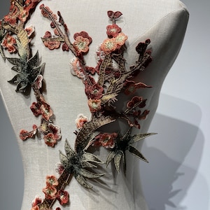 May include: A close-up of a brown and green embroidered applique featuring a floral design with leaves and branches. The applique is on a white fabric background.