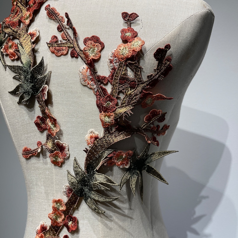 May include: A close-up of a brown and green embroidered applique featuring a floral design with leaves and branches. The applique is on a white fabric background.