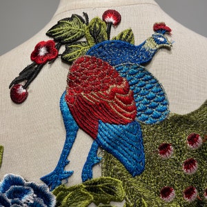 May include: Embroidered peacock patch with vibrant colors. The peacock features a red and gold body, blue wings and tail, and a blue head. Green leaves and red flowers are also embroidered on the patch. The patch is likely for clothing or craft projects.