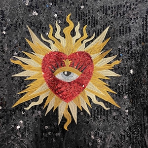 A red heart with a blue eye in the center, surrounded by a gold sunburst design on a black sequined background.