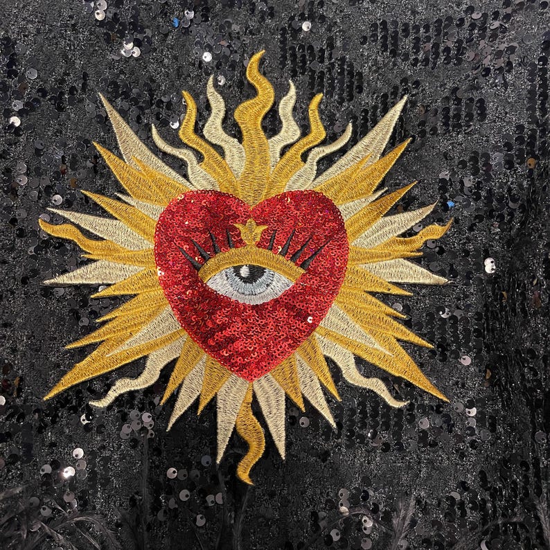 A red heart with a blue eye in the center, surrounded by a gold sunburst design on a black sequined background.