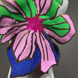 May include: A large pink, green, and blue sequined flower applique. The flower has a central pink circle and multiple petals.