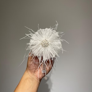 May include: A white feather flower hair clip with a pearl and rhinestone embellishment. The clip has a dangling pearl charm.