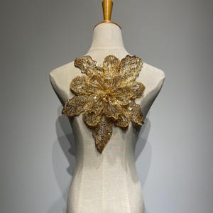 May include: A gold flower-shaped embellishment with sequins and beads. The embellishment is on a white mannequin.