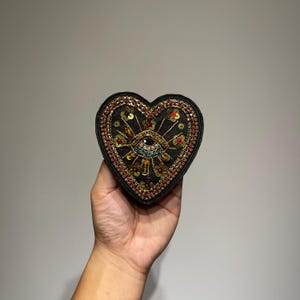 Può includere: Una toppa a forma di cuore nera con un disegno a occhio, ornata di perline e paillettes dorate, rosse e blu. L'occhio è al centro, con raggi di sole e dettagli floreali. La toppa è tenuta su uno sfondo neutro.