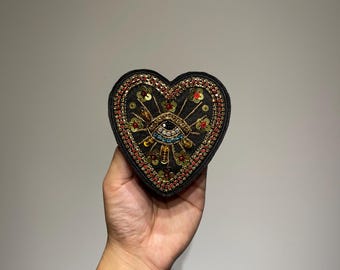 Può includere: Una toppa a forma di cuore nera con un disegno a occhio, ornata di perline e paillettes dorate, rosse e blu. L'occhio è al centro, con raggi di sole e dettagli floreali. La toppa è tenuta su uno sfondo neutro.