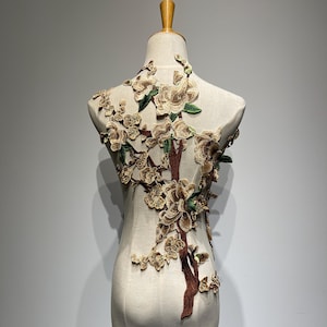 Puede incluir: Una aplicación floral bordada en marrón, beige y verde que representa una rama de árbol con flores y hojas. La aplicación está unida a un maniquí de tela blanca.