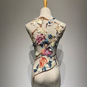 Può includere: Un manichino bianco con un ricamo floreale. Il ricamo presenta un uccello, fiori e foglie in tonalità di rosa, arancio, giallo e blu.