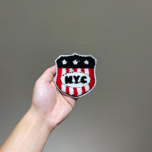 May include: A red, white, and blue embroidered patch with a shield shape and the text "NYC" in the center. The patch has a black outline and five white stars in the top portion of the shield.