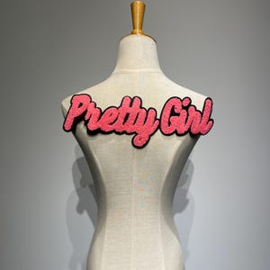 Può includere: Un toppa di feltro rosa e nero con il testo "Pretty Girl" su un manichino bianco.