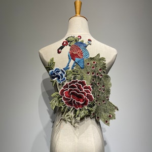 May include: An embroidered peacock patch with a vibrant design. The patch features a peacock with blue and red plumage, surrounded by a large red peony, blue flower, and green leaves. The patch is displayed on a mannequin.