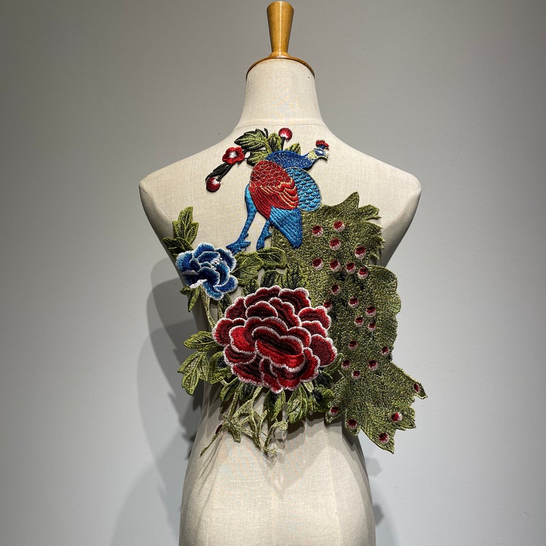 May include: An embroidered peacock patch with a vibrant design. The patch features a peacock with blue and red plumage, surrounded by a large red peony, blue flower, and green leaves. The patch is displayed on a mannequin.