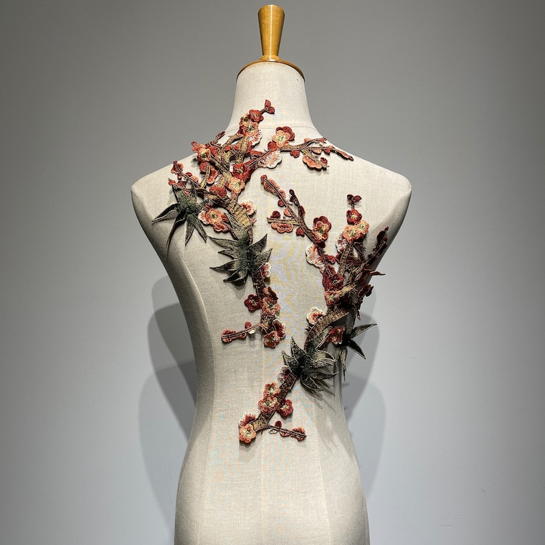 May include: A brown and green embroidered applique featuring a floral design with delicate flowers and leaves. The applique is displayed on a white mannequin.
