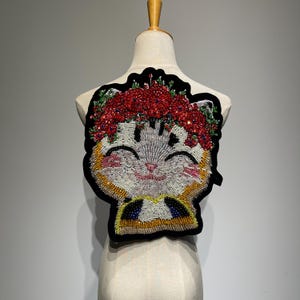 May include: A beaded cat applique with a red floral crown. The cat's face is white, gray, and pink, with a smiling expression. The applique is outlined in black and has yellow and blue accents.