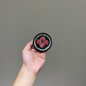 May include: A black and white round patch with a red flower design in the center. The flower has a silver button in the middle.