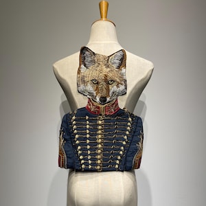 Può includere: Toppa ricamata raffigurante la testa di una volpe su una giacca militare. La giacca è blu navy con bottoni dorati e dettagli rossi. La volpe ha una pelliccia dettagliata marrone e bianca, con un'espressione seria.