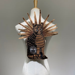 Peut inclure: Une applique brodée de chat marron et noir avec un design de soleil en sequins dorés. L'applique est montée sur un mannequin en tissu blanc.