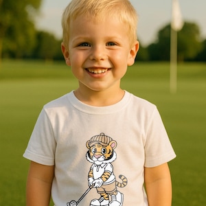 Könnte beinhalten: Ein junges Kind trägt ein cremefarbenes T-Shirt mit einem Cartoon-Tiger, der Golf spielt. Der Tiger trägt eine Golfkappe, ein Poloshirt und Shorts. Das Kind lächelt auf einem grünen Golfplatz.
