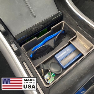May include: A gray plastic organizer tray with compartments for a phone, keys, sunglasses, and other small items. The tray is designed to fit in the center console of a Tesla Model 3 or Model Y. The tray is made in the USA.