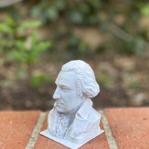 May include: A white 3D printed bust of a historical figure with a textured surface. The bust is on a brick surface.
