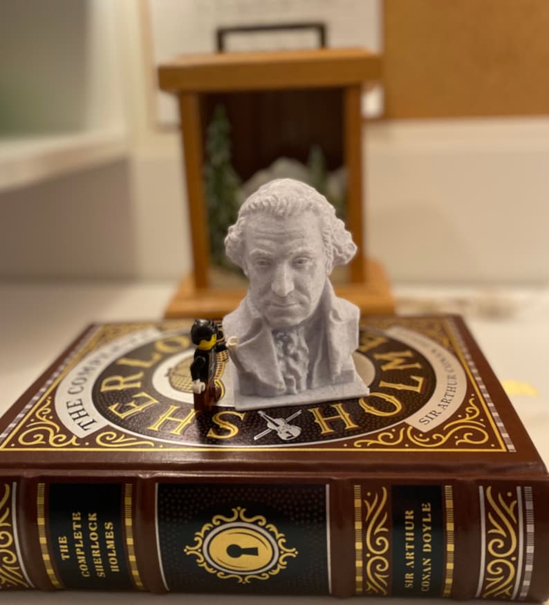 May include: A white bust of a man with a powdered wig, sitting on a book titled "The Complete Sherlock Holmes" by Sir Arthur Conan Doyle. A small black and yellow figurine stands next to the bust.