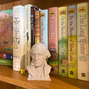 May include: A bookshelf with a variety of books, including the Little House on the Prairie series by Laura Ingalls Wilder. A bust of George Washington is on the shelf in front of the books.