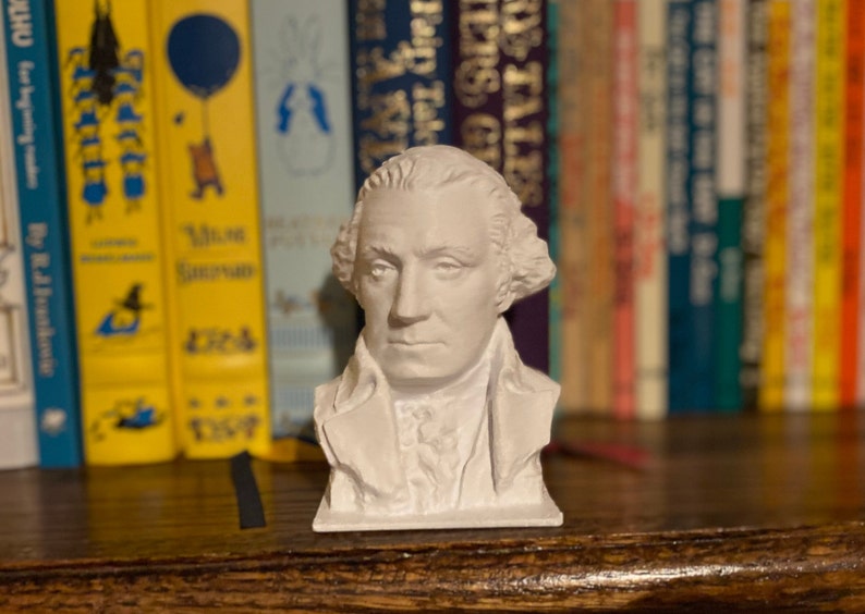 May include: A white bust sculpture of George Washington, the first president of the United States. The bust is on a wooden shelf in front of a bookshelf filled with colorful books.