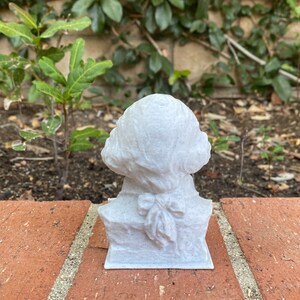 May include: A white bust sculpture of a person with a bow tie. The sculpture is made of a textured material and is sitting on a brick surface.