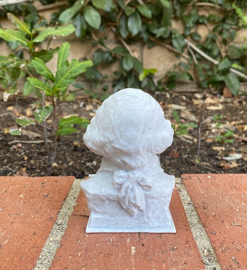 May include: A white bust sculpture of a person with a bow tie. The sculpture is made of a textured material and is sitting on a brick surface.