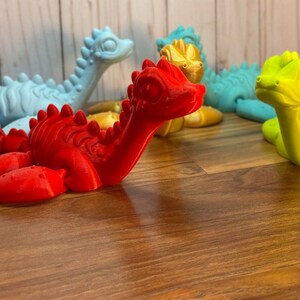 May include: Four small dinosaur figurines in different colors: light blue, gold, red, and green. The dinosaurs are posed in a playful manner, with their heads tilted and their mouths slightly open.