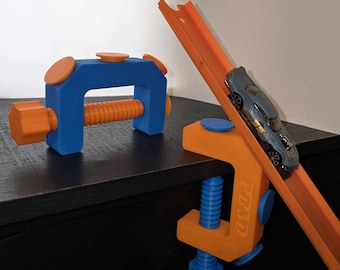 May include: Two orange and blue 3D printed clamps with a screw mechanism. One clamp is holding a section of orange Hot Wheels track with a blue toy car on it.