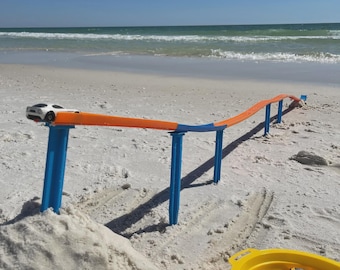 May include: A toy car track set up on a sandy beach with a white and black toy car at the start. The track is orange and blue, supported by blue posts. A yellow toy bin with toy cars is in the foreground, with the ocean in the background.