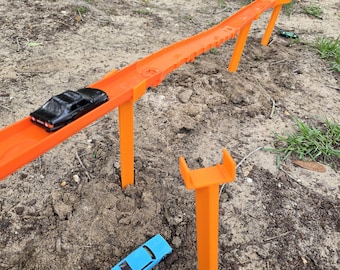 May include: An orange toy race track with a black toy car on it. A blue toy car is on the ground below the track. The track is supported by orange pillars and is set up outdoors on dirt and grass.